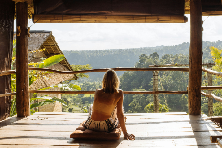 Une personne assise sur une plateforme en bois, regardant un paysage verdoyant de collines et d'arbres à Bali, illuminé par le soleil.